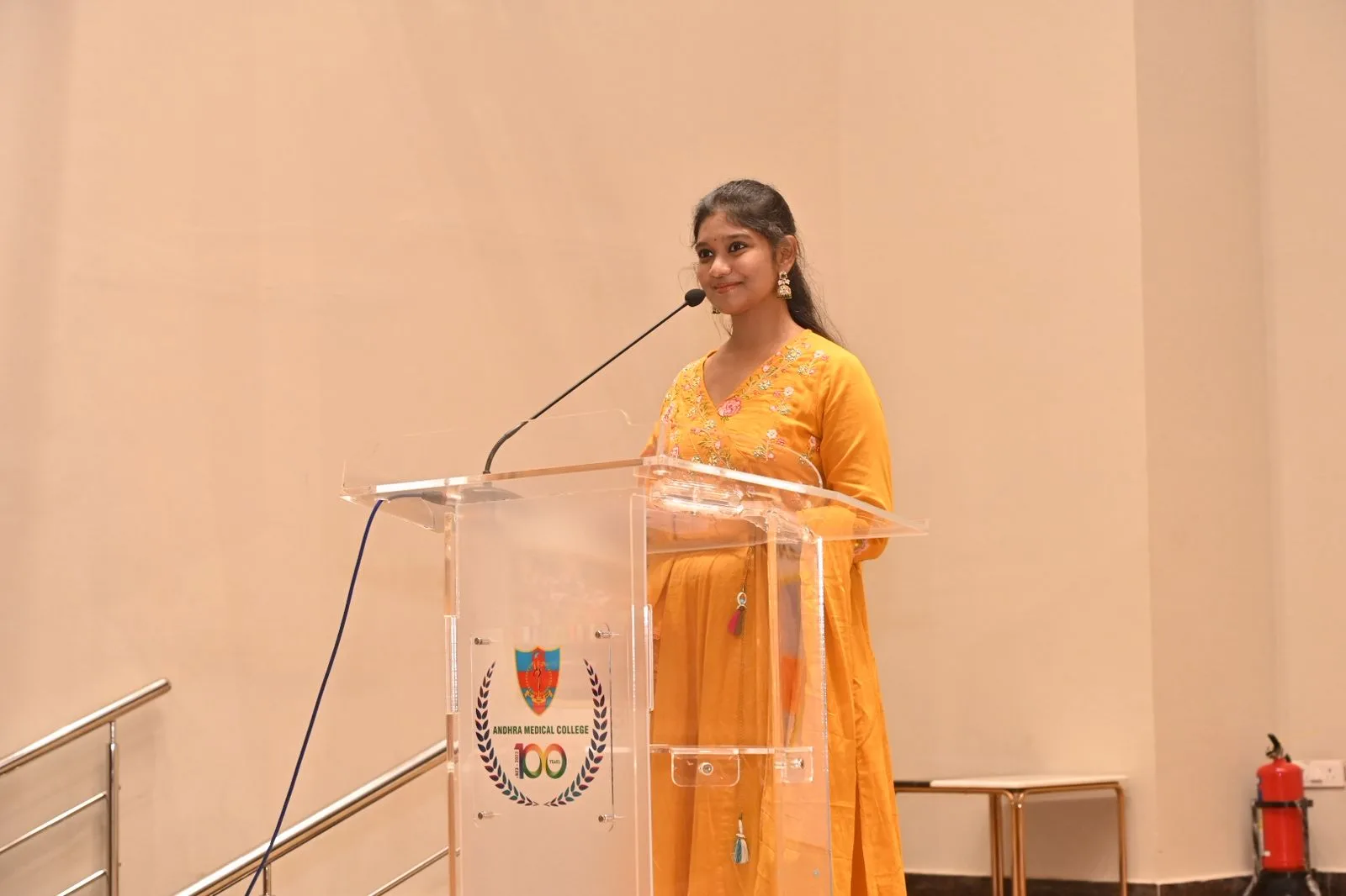 Rashmitha is standing in front of a clear podium with a microphone, wearing a yellow dress and posing for a photo in a beige room.