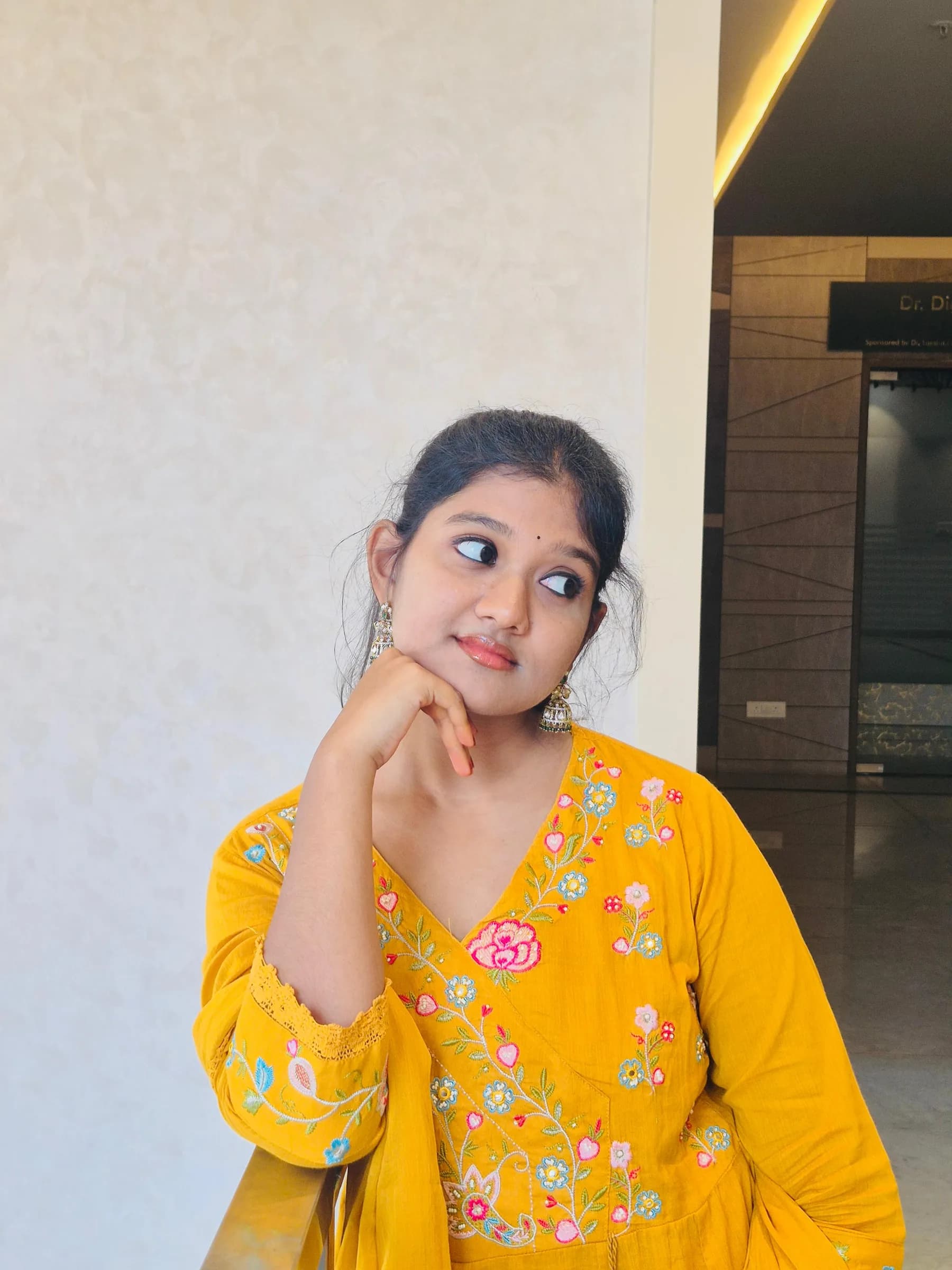 Rashmitha is wearing a yellow dress with floral embroidery, posing in front of a white wall and a doorway, with her hand under her chin.
