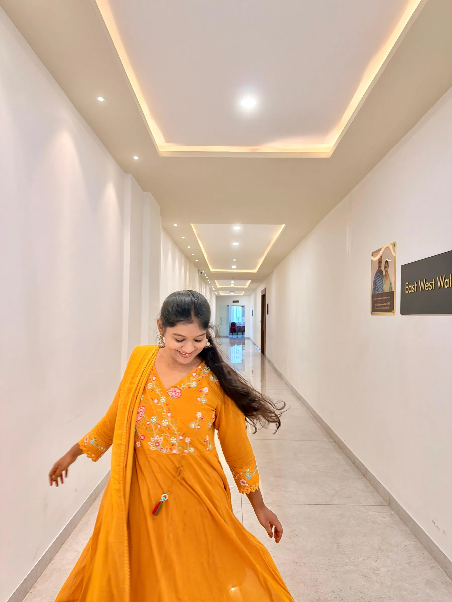 Rashmitha, a model and actress, is wearing a yellow dress and standing in a hallway with white walls and a sign that says East West Walk in a photo