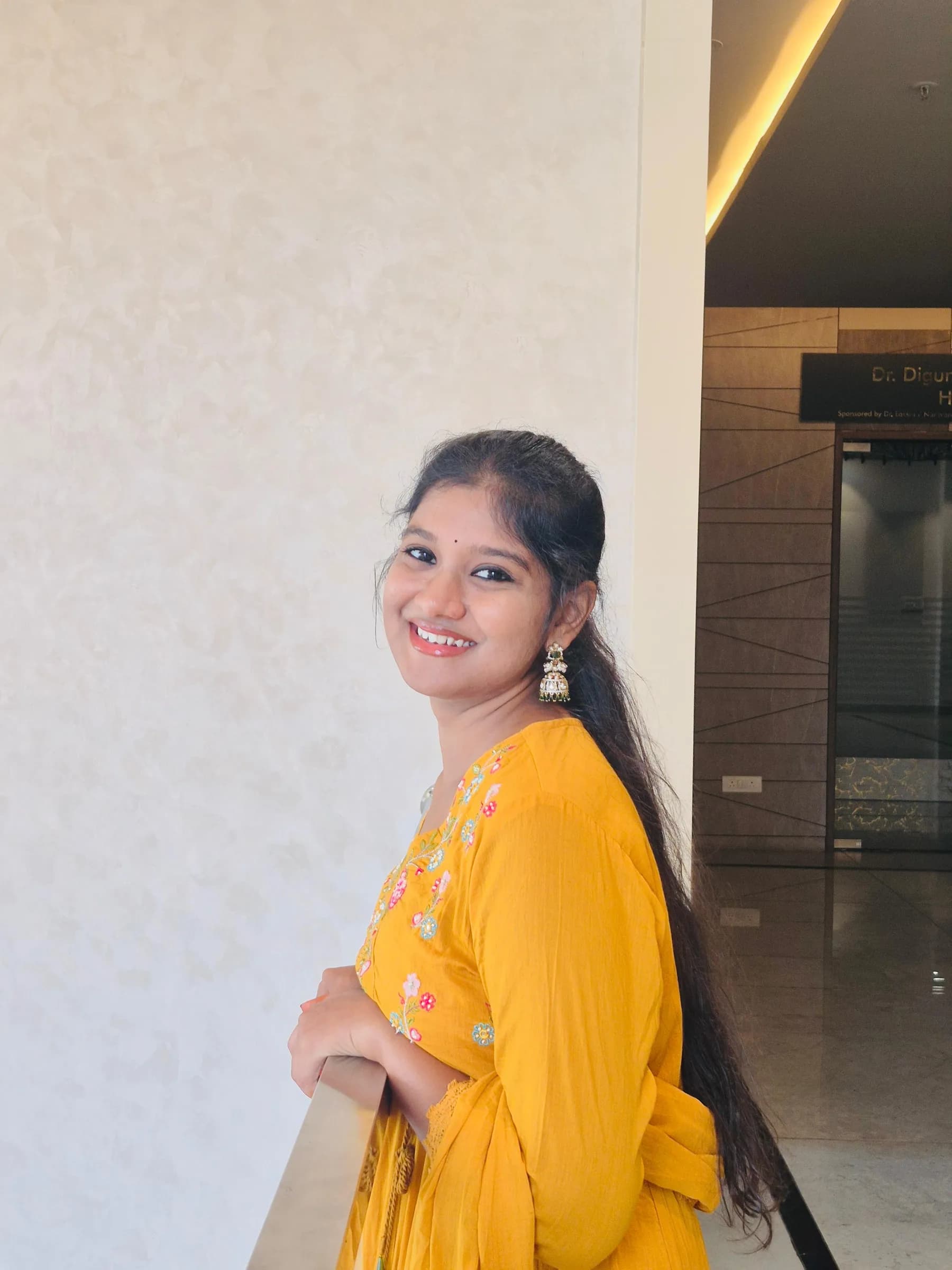 Rashmitha is wearing a yellow dress with floral embroidery, posing in front of a white wall and a glass door,