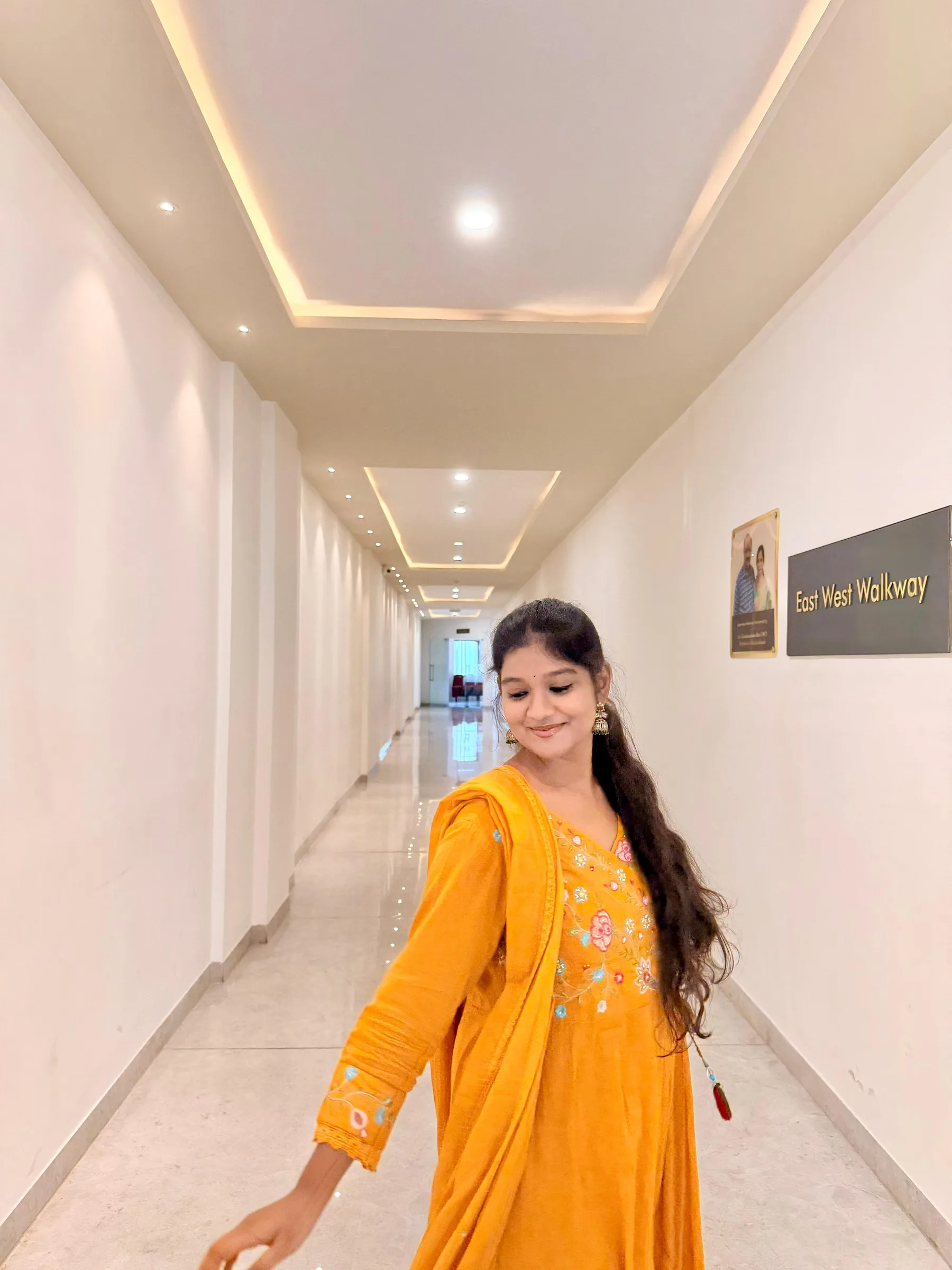 Rashmitha, a model and actress, is wearing a yellow dress with a matching dupatta, posing in a long, white hallway with a sign that reads East West