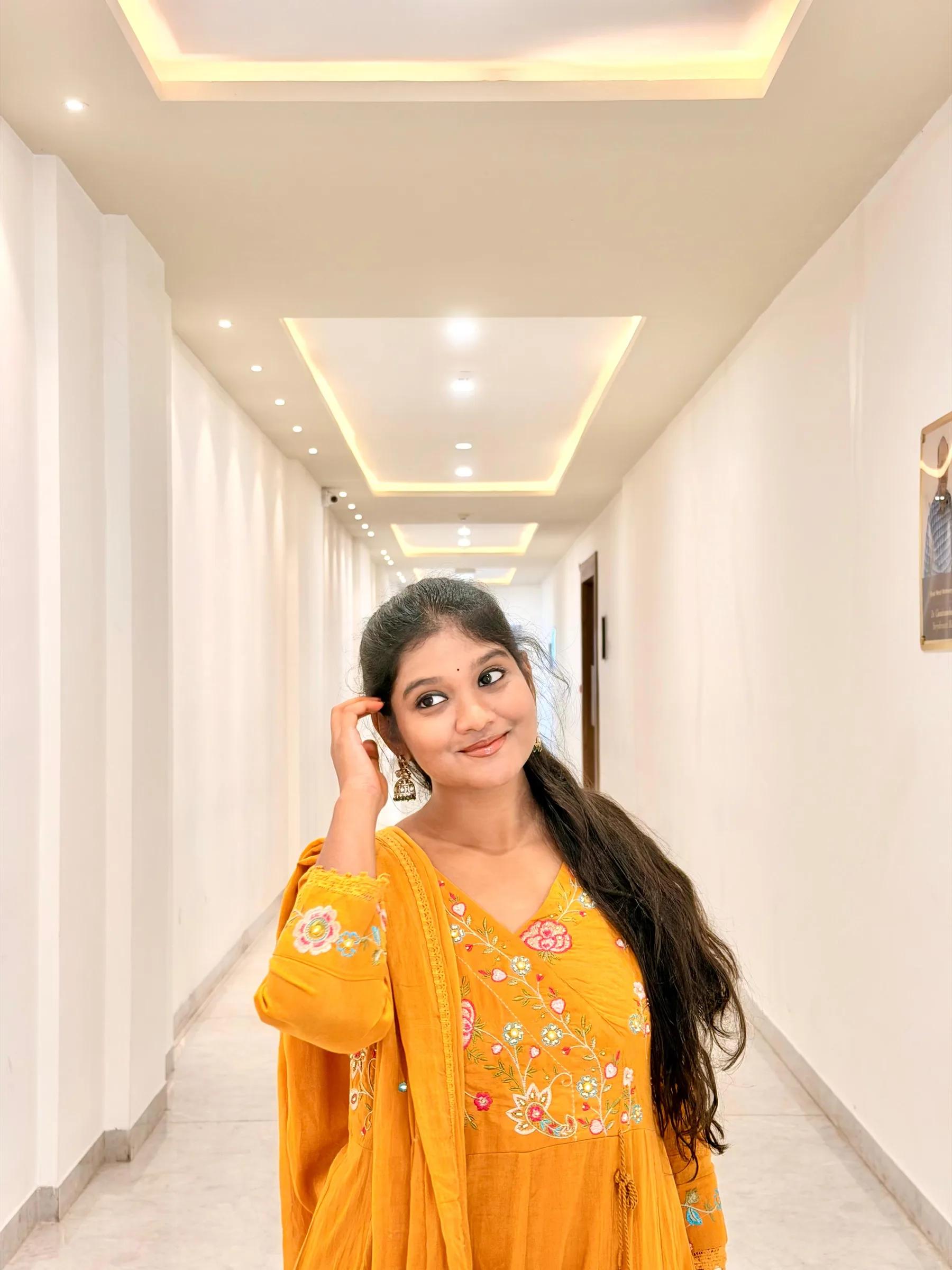 Rashmitha, a model and actress, is dressed in a vibrant yellow outfit with intricate floral embroidery, posing in a long, white hallway with recessed